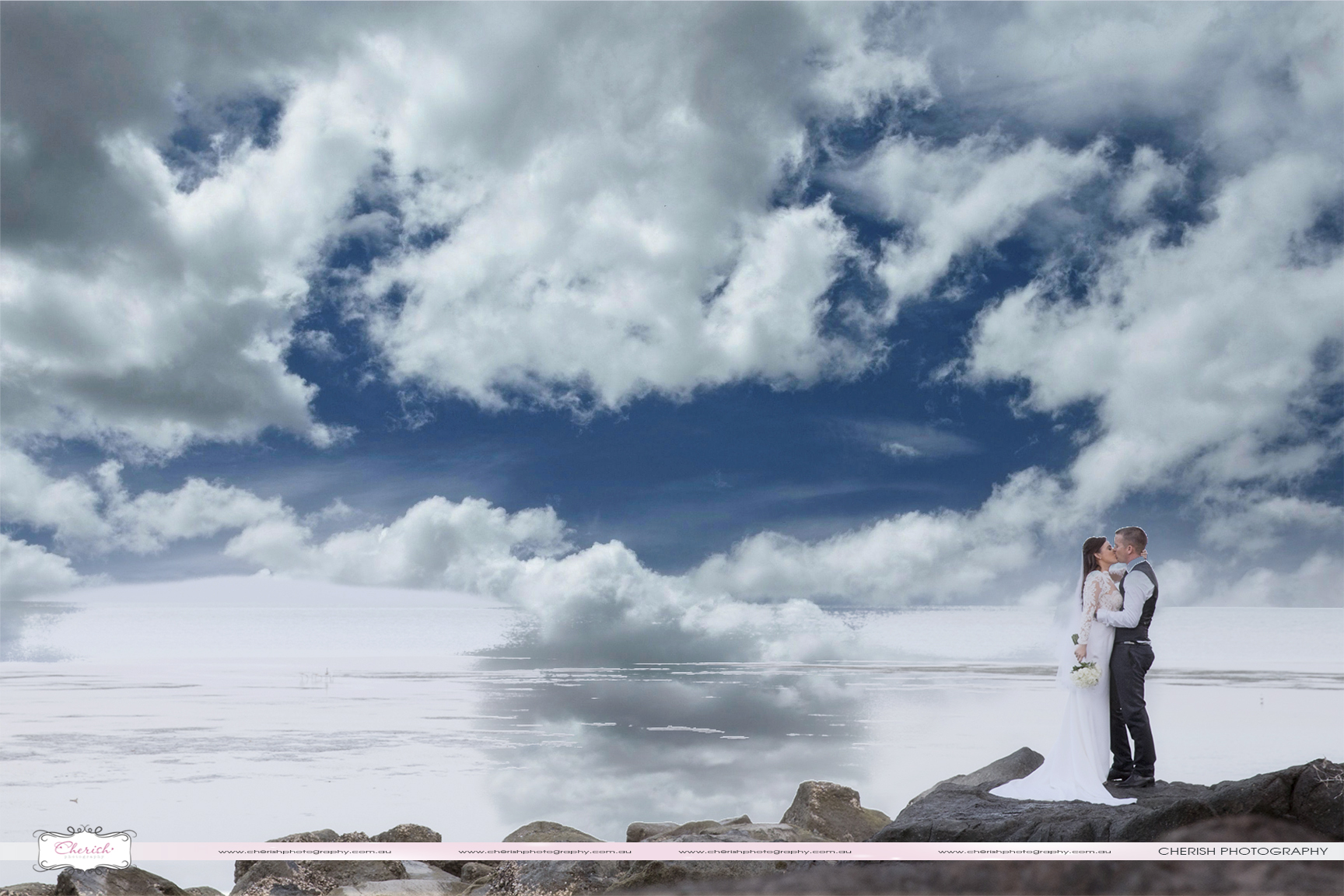 A bride and groom kiss on the beach rocks Villa Botancia with striking clouds.